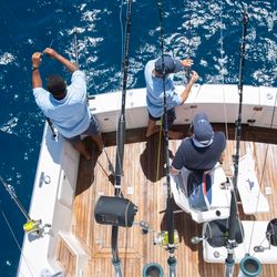 A bird's-eye view of the tail end of a fishing boat with 3 people preparing tackle and bait on large fishing lines.