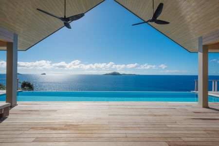 An expansive deck leading out to a wide infinity pool of a residence at Kokomo Island Fiji. Beyond the pool, the calm ocean extends out from below to the horizon, with smaller islands visible in the distance.