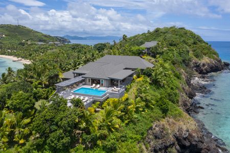 An aerial view of a residence high atop Kokomo Island Fiji, with the residence surrounded lush greenery and its several deck areas and private pool gleaming in sunlight.