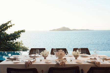 A long dining table elegantly set for a sunset meal on the deck of a private residence at Kokomo Island Fiji. A white coral centrepiece sits between other locally-sourced decorations. The residence’s infitity pool can be seen behind the table setting, past which the ocean extends to the horizon as the sun begins setting.