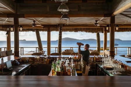 A bartender shaking a cocktail shaker at one of Kokomo Island Fiji’s bars, as the sun begins setting in the distance.