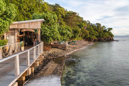 The oceanfront boardwalk leading to Kokomo Island Fiji’s “Walker d'Plank” waterfront restaurant. The restaurant’s elevated deck is seen surrounded by lush greenery as the ocean gently laps the shore beneath.