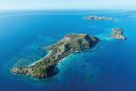 An aerial view of Kokomo Island Fiji surrounded by calm blue waters and reefs. Two small lush islands are seen nearby.