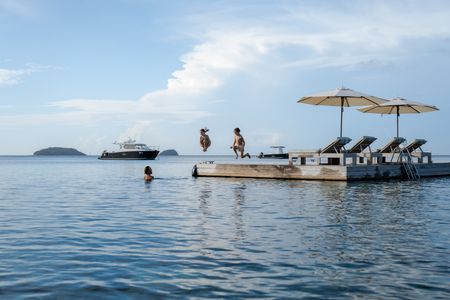 Two children jumping off a floating pontoon into the water, towards their waiting parent. Some of Kokomo Island Fiji’s boats are anchored in the distance.