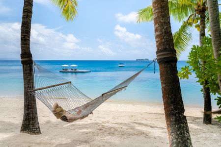 A guest relaxing in a hammock suspended between two palm trees on the beach at Kokomo Island Fiji. In the distance the wide, calm ocean extends to the horizon, with a floating pontoon with several sun lounges anchored just off the shore.
