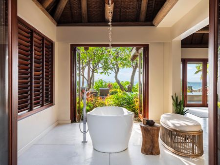 A large free-standing bathtub in a villa at Kokomo Island Fiji, with bay doors open past it showing the villa’s lush private garden.