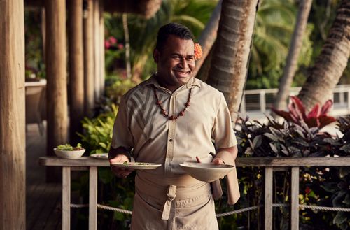 A smiling waiter at Kokomo Private Island Fiji carrying a two meals to a table.
