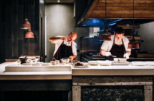 Two chefs at Kokomo Private Island Fiji preparing various dishes. One is pouring seasoning into a bowl while the other works to place food on several plates.