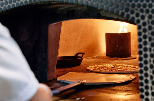 A Kokomo Island chef preparing pizza in a wood-fired oven, with a pizza on a peel and flames in the background