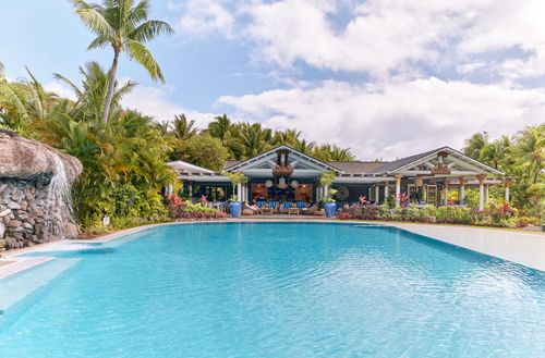 The Kokocabana at Kokomo Private Island Fiji seen from across the wide, blue pool. A small waterfall is falling over rocks into the pool, while several deck chairs can be seen at the entrance to the Pool Club building.
