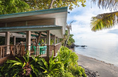 The deck seating area of Walker D’Plank seen from the side, extending out from lush greenery to the sandy shore alongside calm ocean waters.