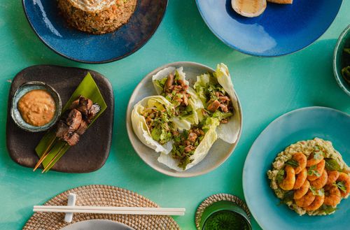Several dishes and drinks served along a vibrant blue-green table seen from above. The dishes include skewers with satay dipping sauce, fried rice, and stuffed lettuce leaves.