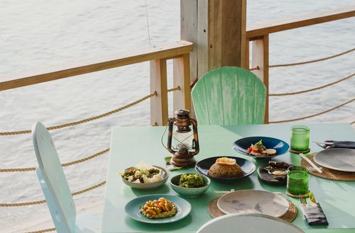 Several dishes and drinks served on a coastal wooden table alongside the edge of the Walker D’Plank dining deck area, with white sand seen directly beyond the deck railing.