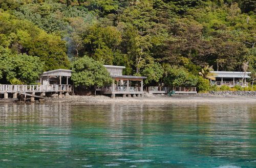 Walker D’Plank seen from out on the ocean, showing the dining area nested amongst lush tropical greenery while its deck extends out over the sand.