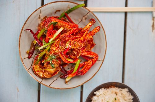 A bright red seafood dish mixed with greens served alongside a bowl of white rice set on a wooden table viewed from above.
